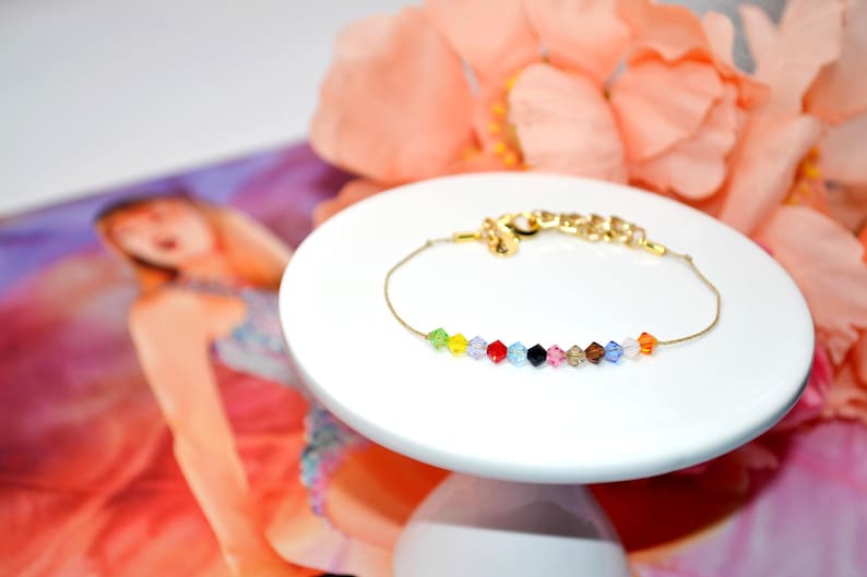A Taylor Swift Eras inspired bracelet displayed on a white pedestal with peach flowers in the background and the Taylor Swift Eras Tour book. The bracelet features a thin gold cord and 12 colorful crystal beads, each representing an era: green (Debut), yellow (Fearless), purple (Speak Now), red (Red), light blue (1989), black (Reputation), pink (Lover), tan (Folklore), brown (Evermore), dark blue (Midnights), white (The Tortured Poets Department), and orange (The Life of a Showgirl).