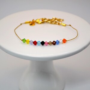 A Taylor Swift Eras inspired bracelet displayed on a white pedestal stand. The bracelet has a thin gold cord with 12 crystal beads, each representing one of Taylor Swift’s eras. The beads are arranged in order: green (Debut), yellow (Fearless), purple (Speak Now), red (Red), light blue (1989), black (Reputation), pink (Lover), tan (Folklore), brown (Evermore), dark blue (Midnights), white (The Tortured Poet's Department), and orange (The Life of a Showgirl).