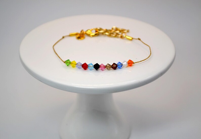 A Taylor Swift Eras inspired bracelet displayed on a white pedestal stand. The bracelet has a thin gold cord with 12 crystal beads, each representing one of Taylor Swift’s eras. The beads are arranged in order: green (Debut), yellow (Fearless), purple (Speak Now), red (Red), light blue (1989), black (Reputation), pink (Lover), tan (Folklore), brown (Evermore), dark blue (Midnights), white (The Tortured Poet's Department), and orange (The Life of a Showgirl).