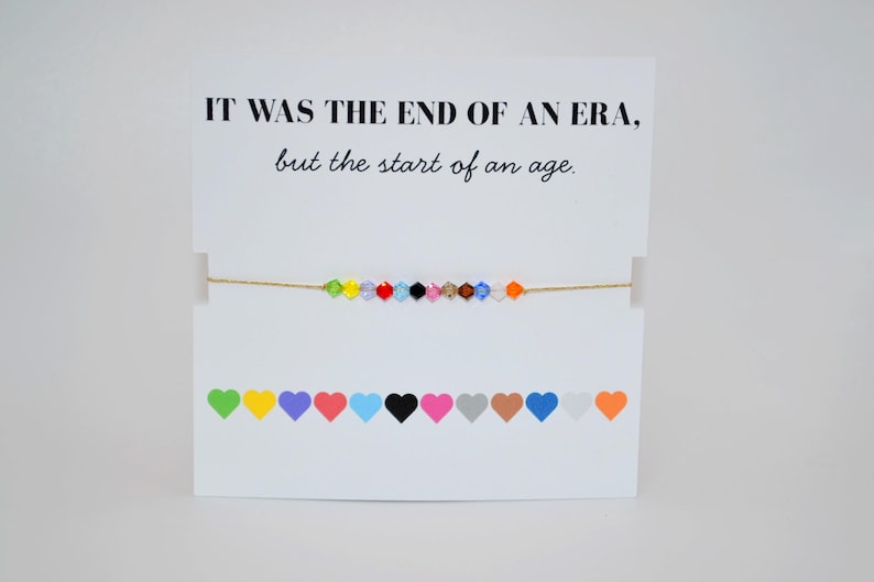 A white card displays a bracelet with colorful crystal beads representing Taylor Swift’s Eras. The top text reads, “IT WAS THE END OF AN ERA, but the start of an age.” Below the bracelet are 12 hearts in different colors, each symbolizing one of Taylor Swift’s musical eras. The bracelet itself features matching multicolored beads strung on a thin gold cord.