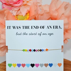 A Taylor Swift Eras bracelet on a white card with peach flowers in the background. The card says, “IT WAS THE END OF AN ERA, but the start of an age.” The gold cord bracelet has 12 colorful beads, each symbolizing an era: Debut (green), Fearless (yellow), Speak Now (purple), Red (red), 1989 (blue), Reputation (black), Lover (pink), Folklore (tan), Evermore (brown), Midnights (dark blue), The Tortured Poets Department (white), and The Life of a Showgirl (orange).
