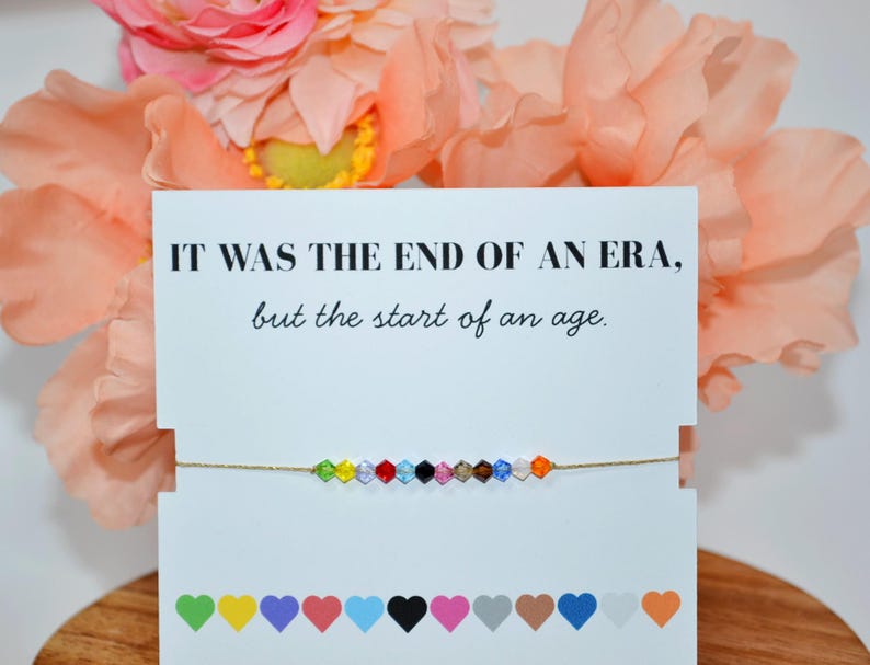 A Taylor Swift Eras bracelet on a white card with peach flowers in the background. The card says, “IT WAS THE END OF AN ERA, but the start of an age.” The gold cord bracelet has 12 colorful beads, each symbolizing an era: Debut (green), Fearless (yellow), Speak Now (purple), Red (red), 1989 (blue), Reputation (black), Lover (pink), Folklore (tan), Evermore (brown), Midnights (dark blue), The Tortured Poets Department (white), and The Life of a Showgirl (orange).