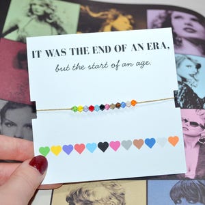 A Taylor Swift Eras bracelet held in front of a collage of her photos. The card reads, “IT WAS THE END OF AN ERA, but the start of an age.” A thin gold cord bracelet with 12 crystal beads represents her eras. At the bottom, 12 hearts show each era: green (Debut), yellow (Fearless), purple (Speak Now), red (Red), light blue (1989), black (Reputation), pink (Lover), tan (Folklore), brown (Evermore), blue (Midnights), white (Tortured Poet's Department), and orange (The Life of a Showgirl).