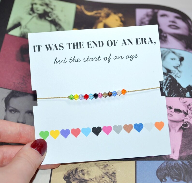 A Taylor Swift Eras bracelet held in front of a collage of her photos. The card reads, “IT WAS THE END OF AN ERA, but the start of an age.” A thin gold cord bracelet with 12 crystal beads represents her eras. At the bottom, 12 hearts show each era: green (Debut), yellow (Fearless), purple (Speak Now), red (Red), light blue (1989), black (Reputation), pink (Lover), tan (Folklore), brown (Evermore), blue (Midnights), white (Tortured Poet's Department), and orange (The Life of a Showgirl).