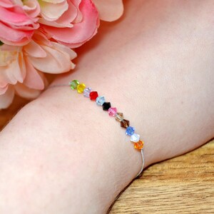A Taylor Swift Eras inspired bracelet worn on a wrist, with peach and pink flowers in the background. The bracelet has a thin silver cord and 12 colorful crystal beads, each symbolizing a Taylor Swift era: green (Debut), yellow (Fearless), purple (Speak Now), red (Red), light blue (1989), black (Reputation), pink (Lover), tan (Folklore), brown (Evermore), dark blue (Midnights), white (The Tortured Poets Department), and orange (The Life of a Showgirl).