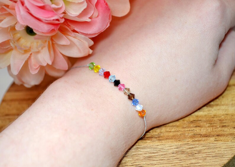 A Taylor Swift Eras inspired bracelet worn on a wrist, with peach and pink flowers in the background. The bracelet has a thin silver cord and 12 colorful crystal beads, each symbolizing a Taylor Swift era: green (Debut), yellow (Fearless), purple (Speak Now), red (Red), light blue (1989), black (Reputation), pink (Lover), tan (Folklore), brown (Evermore), dark blue (Midnights), white (The Tortured Poets Department), and orange (The Life of a Showgirl).