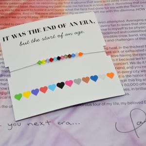 A Taylor Swift Eras inspired bracelet displayed on a white card with the words “IT WAS THE END OF AN ERA, but the start of an age.” The bracelet has 12 colorful crystal beads on a thin silver cord, each symbolizing a Taylor Swift era. At the bottom of the card, 12 matching hearts label each era in colors green, yellow, purple, red, light blue, black, pink, tan, brown, dark blue, white, and orange. The card rests on a page of Taylor Swift’s Eras Tour booklet with her signature at the bottom.