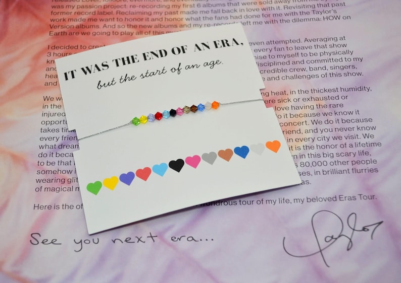 A Taylor Swift Eras inspired bracelet displayed on a white card with the words “IT WAS THE END OF AN ERA, but the start of an age.” The bracelet has 12 colorful crystal beads on a thin silver cord, each symbolizing a Taylor Swift era. At the bottom of the card, 12 matching hearts label each era in colors green, yellow, purple, red, light blue, black, pink, tan, brown, dark blue, white, and orange. The card rests on a page of Taylor Swift’s Eras Tour booklet with her signature at the bottom.