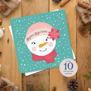 May include: A set of 10 greeting cards featuring a snowman design. The snowman has a pink hat, scarf, and a poinsettia. The card has the words "Happy Holidays" on it, and is set against a teal background with white dots.