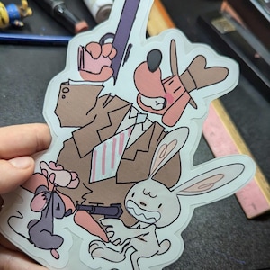 May include: A sticker featuring cartoon characters, including a dog in a fedora, a rabbit, and a mouse, all holding weapons. The characters are set against a brown background with a suit and tie.