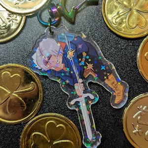 May include: A clear acrylic keychain featuring a character with white hair and a sword, surrounded by gold coins with shamrock designs. The keychain has a holographic effect. The coins are scattered around the keychain.