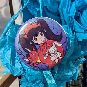 May include: A round button featuring a cartoon character with black hair, a red dress, and a devil and bunny. The button has a blue background with a dotted pattern and is hanging from a blue ribbon.