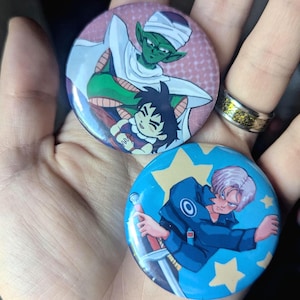 May include: Two round pinback buttons featuring anime characters. One button has a pink background with a green and white character and a black-haired character. The other button is blue with a character holding a sword.