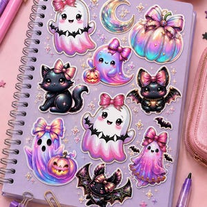 Op de afbeelding: Een lichtpaars spiraalnotitieboek bedekt met diverse stickers van iriserende geesten, zwarte katten, vleermuizen en pompoenen, allemaal versierd met roze strikken. De stickers zijn verspreid over de voorkant van de kaft van het notitieboek.