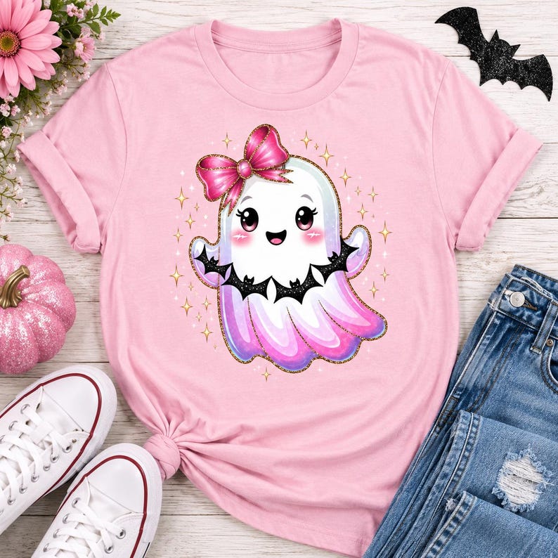 Op de afbeelding: Een lichtroze T-shirt met korte mouwen en een centrale afbeelding van een schattig wit spookje met een roze glitterstrik, roze wangen en zwarte vleugels met vleermuispatroon, omringd door kleine gouden sterretjes op de stof.