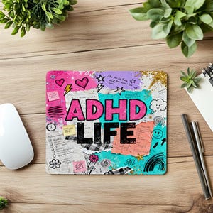 Może przedstawiać: Prostokątna podkładka pod mysz z napisem "ADHD LIFE" w kolorze różowym i czarnym. Projekt zawiera serca, gwiazdy, błyskawice i odręczne notatki na tle kolażu. Podkładka pod mysz leży na drewnianej powierzchni.
