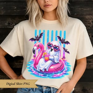 May include: A cream-colored t-shirt featuring a graphic of a white ghost wearing heart-shaped sunglasses, sitting in a pink flamingo pool float while holding a tropical drink. Three small black bats fly above, with palm trees and blue water.