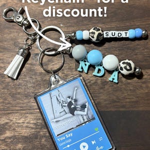May include: A keychain bundle featuring a rectangular acrylic music player charm with a blue interface, the text "You Say" by Lauren Daigle, and a dancer image. It includes beaded strands with light blue, grey, and leopard print beads, letter beads spelling "SUDT" and "NDA", and a white tassel.