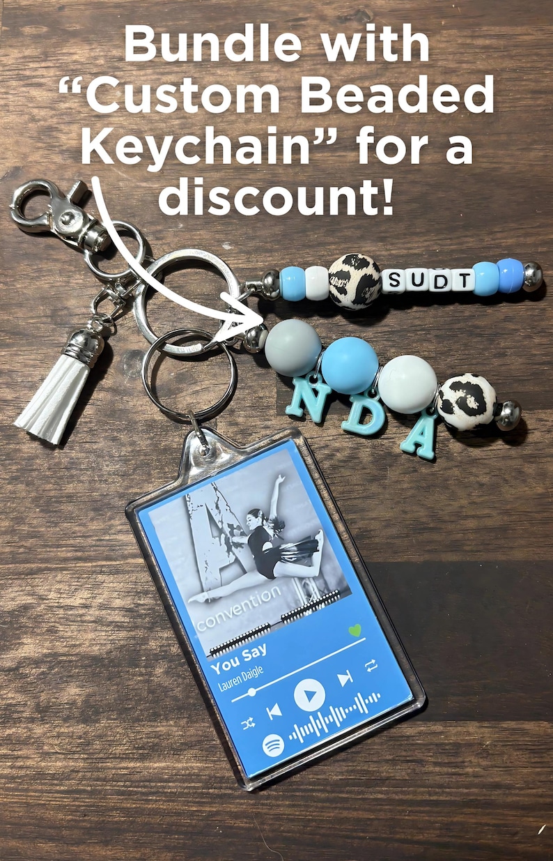 May include: A keychain bundle featuring a rectangular acrylic music player charm with a blue interface, the text "You Say" by Lauren Daigle, and a dancer image. It includes beaded strands with light blue, grey, and leopard print beads, letter beads spelling "SUDT" and "NDA", and a white tassel.