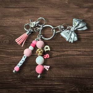 May include: A pink and white beaded keychain with the name "Ella" spelled out in pink letters. The keychain also has a silver bow charm and a pink tassel charm.