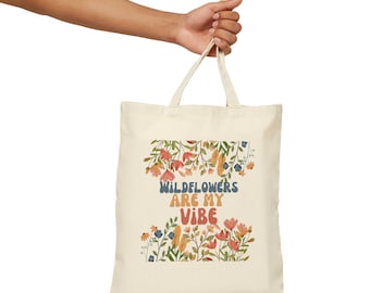 Op de afbeelding: Een witte canvas tote bag met een bloemenontwerp en de tekst "Wildflowers are my vibe".