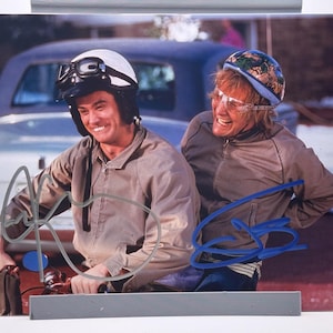 May include: A photograph featuring two men smiling, wearing helmets and jackets. One man wears a white helmet with goggles, the other a blue patterned helmet. Both are in front of a vintage car.