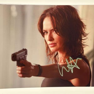 May include: A framed print featuring a woman with brown hair holding a black handgun. The woman is wearing a black top and a black wristband. A green signature is visible on the print.