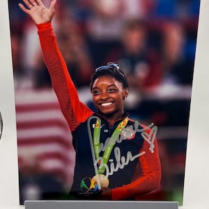 May include: A photograph of a smiling athlete wearing a navy blue and red uniform, holding two gold medals, and raising her right arm in celebration. The athlete's signature is visible on the photo. The background is blurred, suggesting a stadium setting.