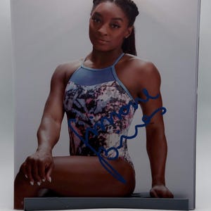 May include: A photograph of a person wearing a blue and patterned leotard with a signature in blue ink. The person is seated, with arms and legs visible. The background is a light gray.