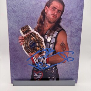 Peut inclure: Photographie encadrée d'un lutteur tenant une ceinture de championnat. Le lutteur porte un gilet noir avec des détails argentés et un tatouage. La ceinture est dorée et blanche avec des accents bleus, et la photo est signée à l'encre bleue.