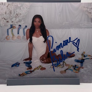 Puede incluir: Fotografía de una mujer con un vestido blanco, sentada sobre una superficie blanca. Se ven varias medallas con cintas azules. La imagen está firmada con tinta azul. Flores blancas decorativas en el fondo.