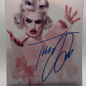 May include: A print featuring a person with dramatic makeup, styled hair, and a pink and white outfit. The print is signed in blue ink. The background has a watercolor effect with pink and white hues. The person is looking directly at the viewer.