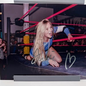 May include: A signed photograph of a female wrestler in a wrestling ring. She is kneeling on the mat, wearing a blue and white outfit, with long blonde hair. The image includes a heart and signature.