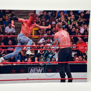 May include: A photograph of a wrestling match in progress. One wrestler is jumping over the ropes, while another stands in the ring. Both wrestlers are covered in what appears to be blood. The crowd is visible in the background.