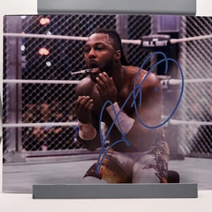 May include: A signed photograph of a wrestler in a wrestling ring. The wrestler is kneeling, holding a syringe to his mouth. The image is signed in blue ink. The background shows a chain-link fence and the words "ALL OUT".