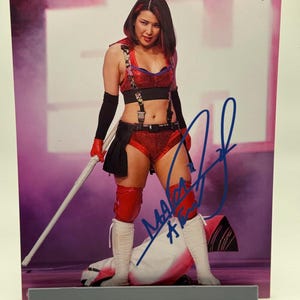 May include: A signed photograph of a wrestler in a red and black outfit, holding a white staff. The wrestler is wearing knee pads and white boots. The background is a blurred pink and white. The signature is in blue ink.