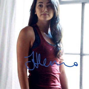 Può includere: Una donna indossa una canotta rossa con una firma blu. Ha lunghi capelli scuri e si trova vicino a una finestra. La canotta è senza maniche e sembra essere fatta di un materiale morbido. L'immagine è ben illuminata.