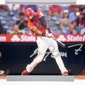 Peut inclure: Photographie signée d'un joueur de baseball en action, en train de frapper la balle. Le joueur porte un uniforme rouge, un pantalon blanc et des chaussures rouges. En arrière-plan, un stade de baseball.