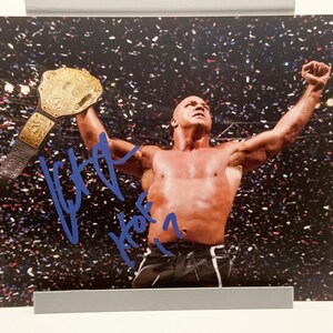 Puede incluir: Fotografía enmarcada de un hombre musculoso sosteniendo un cinturón de campeonato dorado sobre su cabeza. El hombre está sin camisa y lleva pantalones cortos negros. El fondo está lleno de confeti y la foto está firmada con tinta azul.