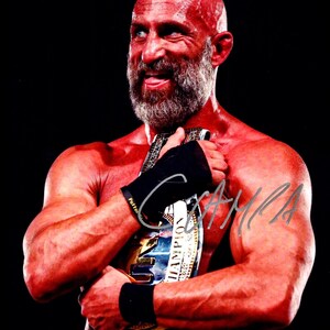 May include: A muscular man with a bald head and a gray beard holds a championship belt. He is wearing black wristbands and has a signature on his arm. The belt is gold and blue with the word "CHAMPION" visible.