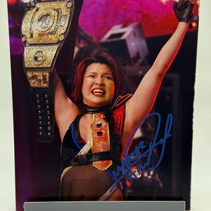May include: A framed photograph of a wrestler holding a championship belt aloft. The wrestler is wearing a black and red wrestling outfit and has red hair. The belt is gold and black.