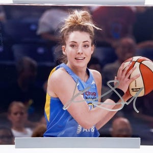May include: A framed photograph of a basketball player holding a basketball. The player is wearing a blue and yellow jersey and has a signature on the photograph. The basketball is orange, white, and brown.