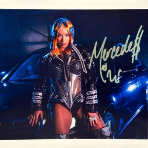 May include: A photograph featuring a person in a futuristic outfit, posing next to a dark blue car. The outfit is black with silver accents. The person's hair is styled in a vibrant mix of orange and blue. The image is signed with the name "Mercedes" in green ink.