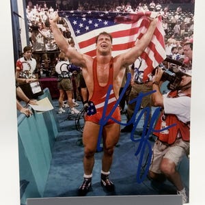 Puede incluir: Fotografía enmarcada de un luchador con un maillot rojo y azul sosteniendo una bandera estadounidense. El luchador sonríe y levanta los brazos en señal de victoria. La foto está firmada con tinta azul.