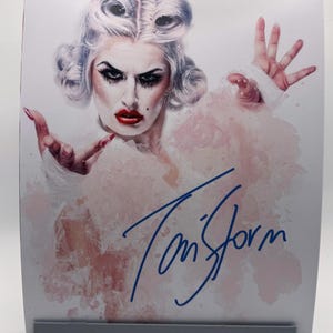 May include: A print featuring a portrait of a person with dramatic makeup, styled hair, and a white fluffy garment. The print is signed in blue ink with the name "Tom Storm". The background has a watercolor effect with pink and red hues.