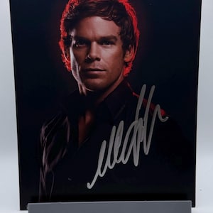 May include: A glossy photograph featuring a man with short brown hair and a serious expression. The background is dark, with a red glow around the head. The photograph is signed in silver ink.