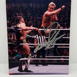 May include: A signed photograph of a wrestling match in progress. Two wrestlers are in the ring, one in mid-air, with a referee in the background. The image is signed in silver ink. The crowd is visible in the background.