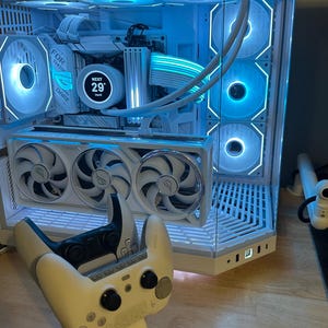 May include: A white gaming PC with blue LED lighting, featuring multiple fans and a clear side panel. A white game controller rests in front of the computer. The PC has the text "FOR THOSE WHO DARE".