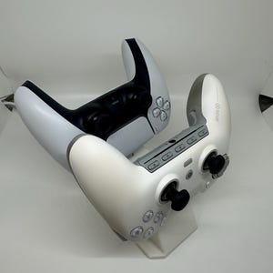 May include: Two white and black game controllers are displayed against a white background. One controller is black and white, while the other is white and grey. The controllers have buttons and joysticks.
