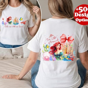 Puede incluir: Camiseta blanca con un gr&aacute;fico colorido de una sirena, un castillo y criaturas marinas. La parte delantera de la camiseta tiene el texto "Tale as old as time". La parte trasera de la camiseta tiene el texto "The Little Mermaid".